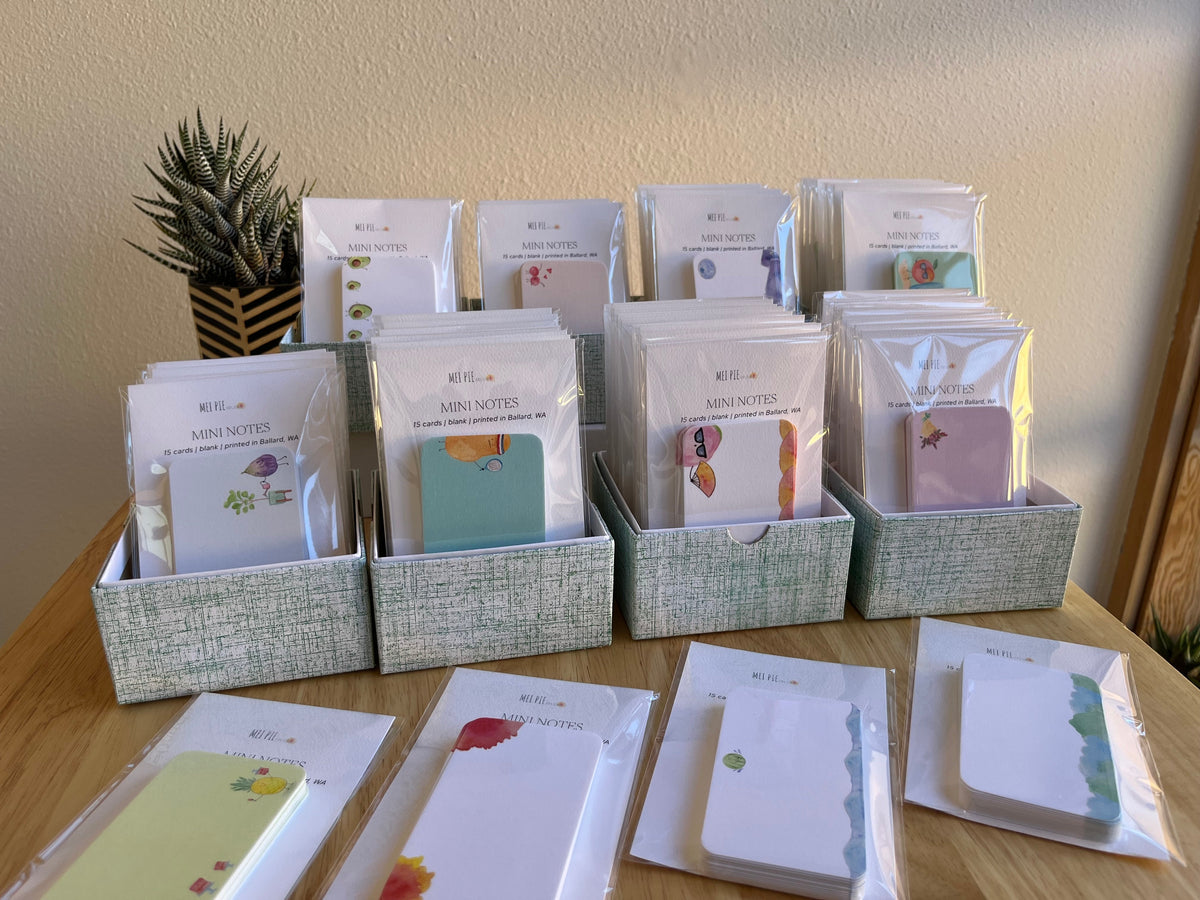 A collection of small stationery items, including cards, mini notes, and small notebooks, displayed in three gray fabric bins on a wooden table against a beige wall.