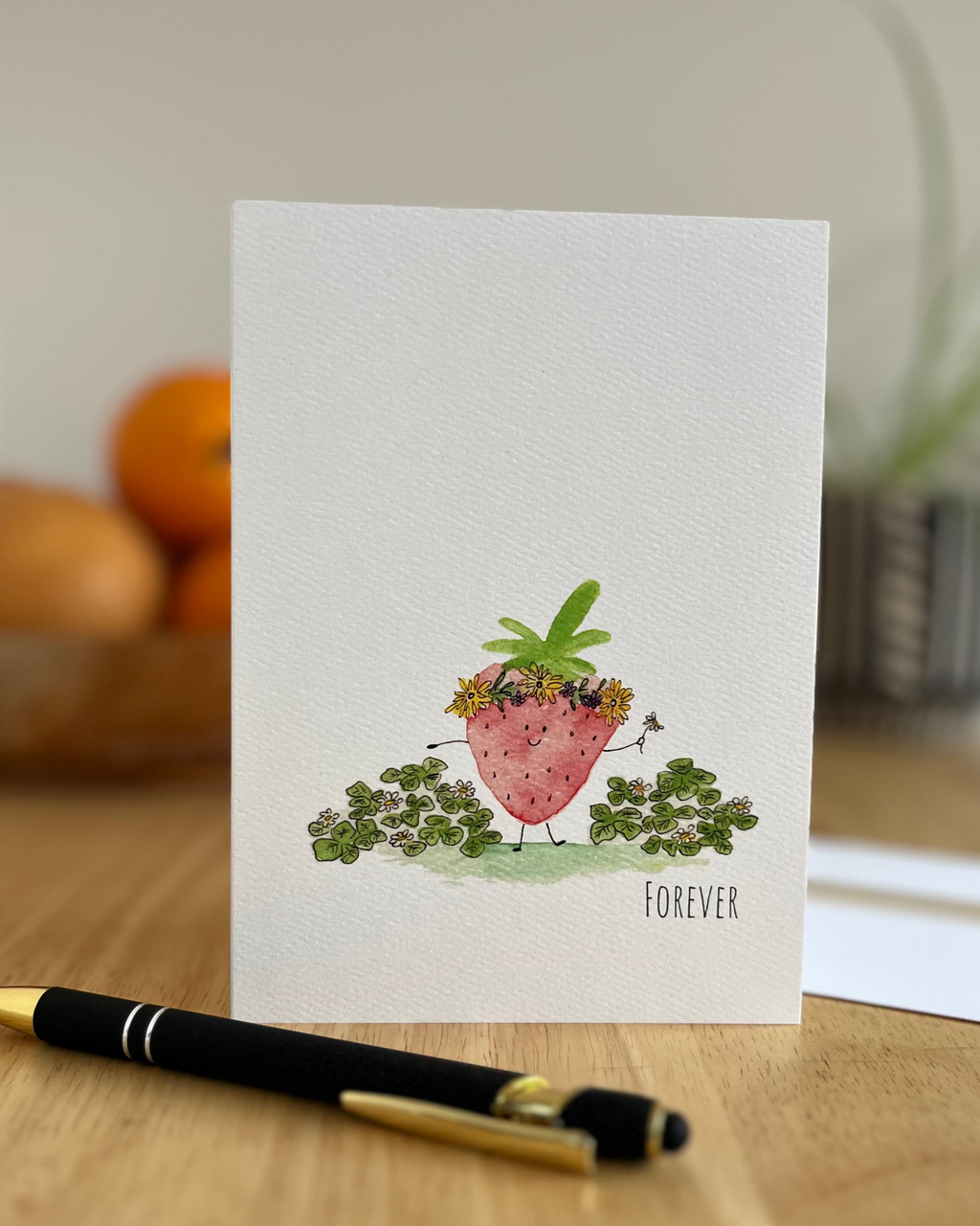 Greeting card with strawberry illustration on a wooden surface with a pen.