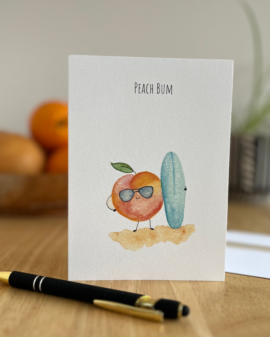 Card with a cartoon peach and cactus character labeled 'Peach Bum' on a wooden surface.
