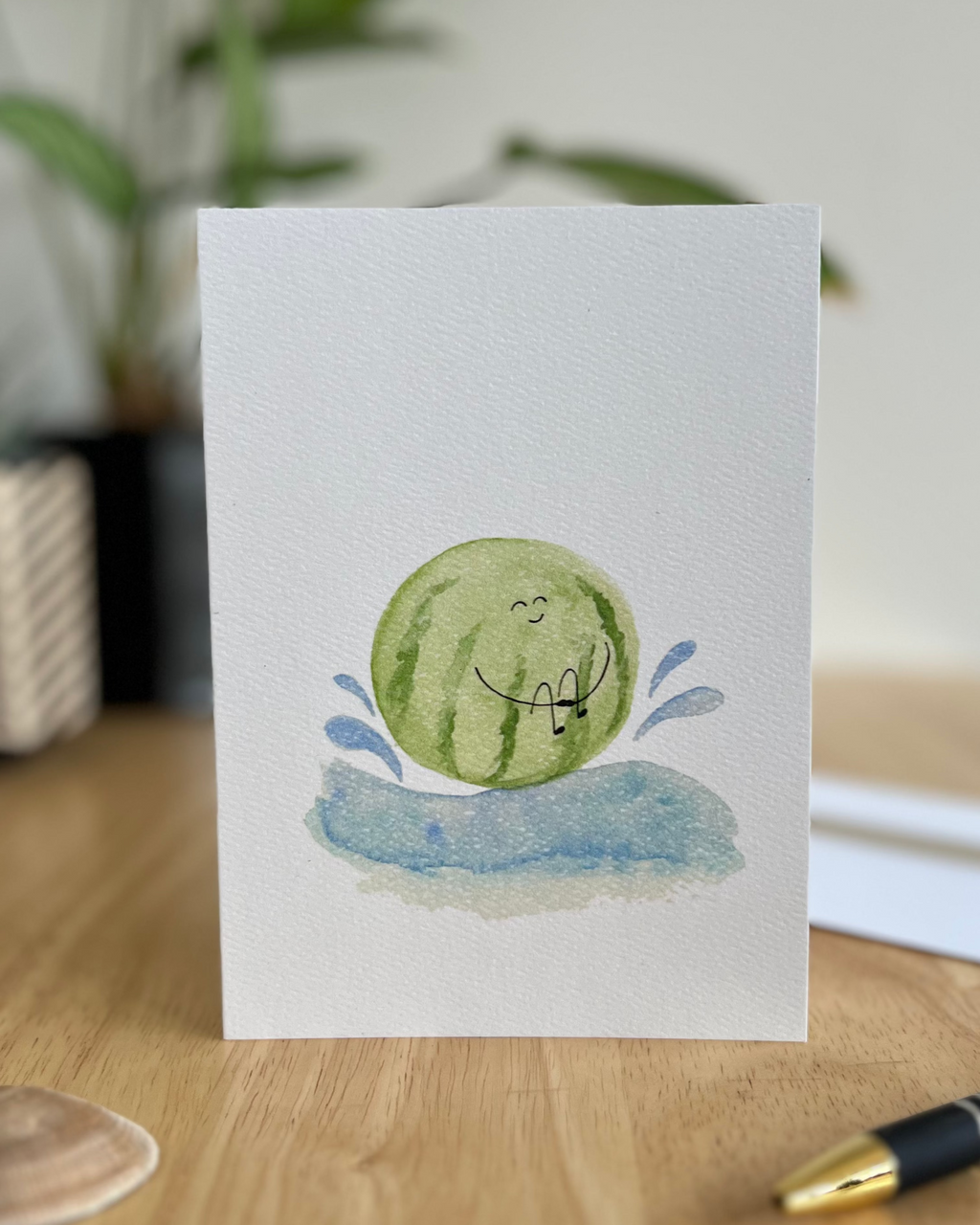 Whimsical watercolor illustration of a round watermelon splashing into blue water with water droplets flying.