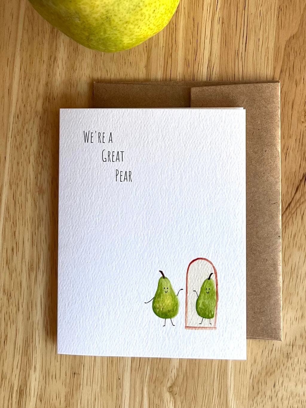 Greeting card with two pears and text 'We're a great pear' on a wooden surface with a pear above the card.