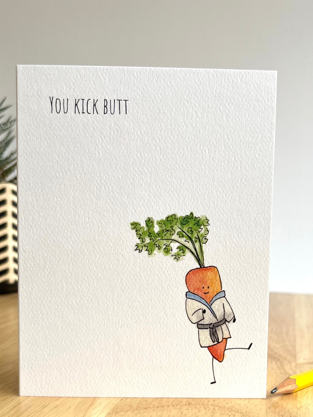 Greeting card with a carrot character in a kimono and text 'You Kick Butt' on a wooden surface.