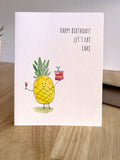 Birthday card with a pineapple holding a cake and candle, on a wooden surface.