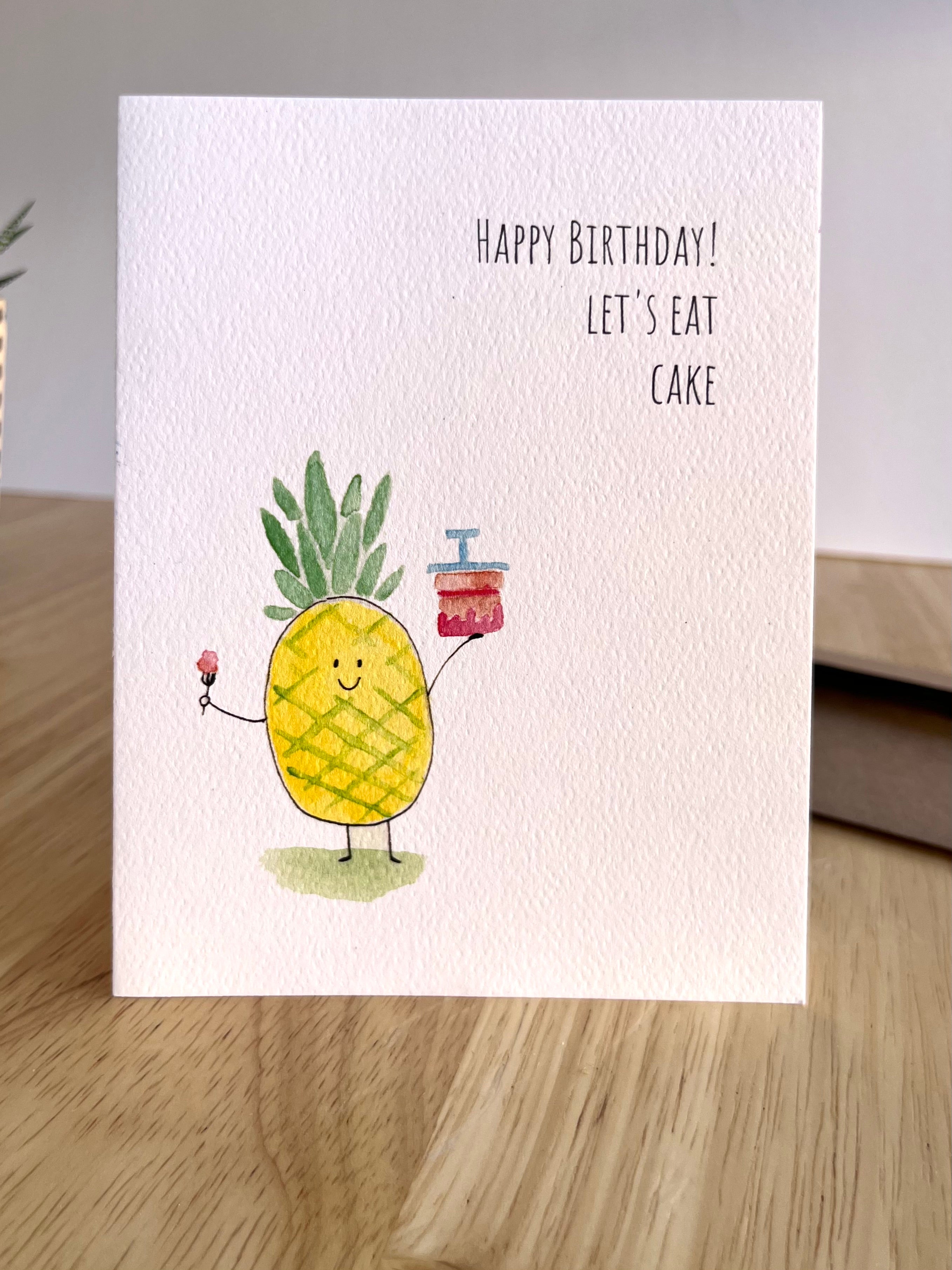 Birthday card with a pineapple holding a cake and candle, on a wooden surface.