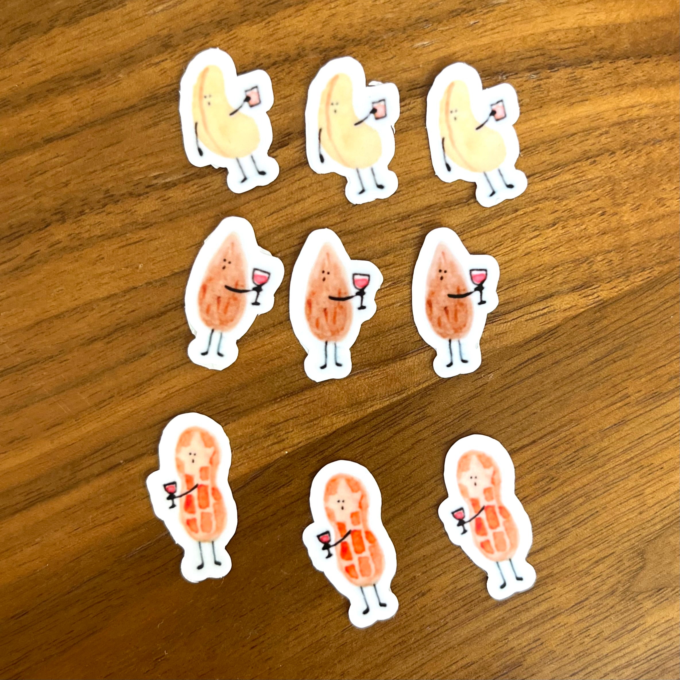 Nine die-cut stickers featuring cute cartoon characters shaped like peanuts and almonds holding wine glasses, arranged on a brown wooden surface.