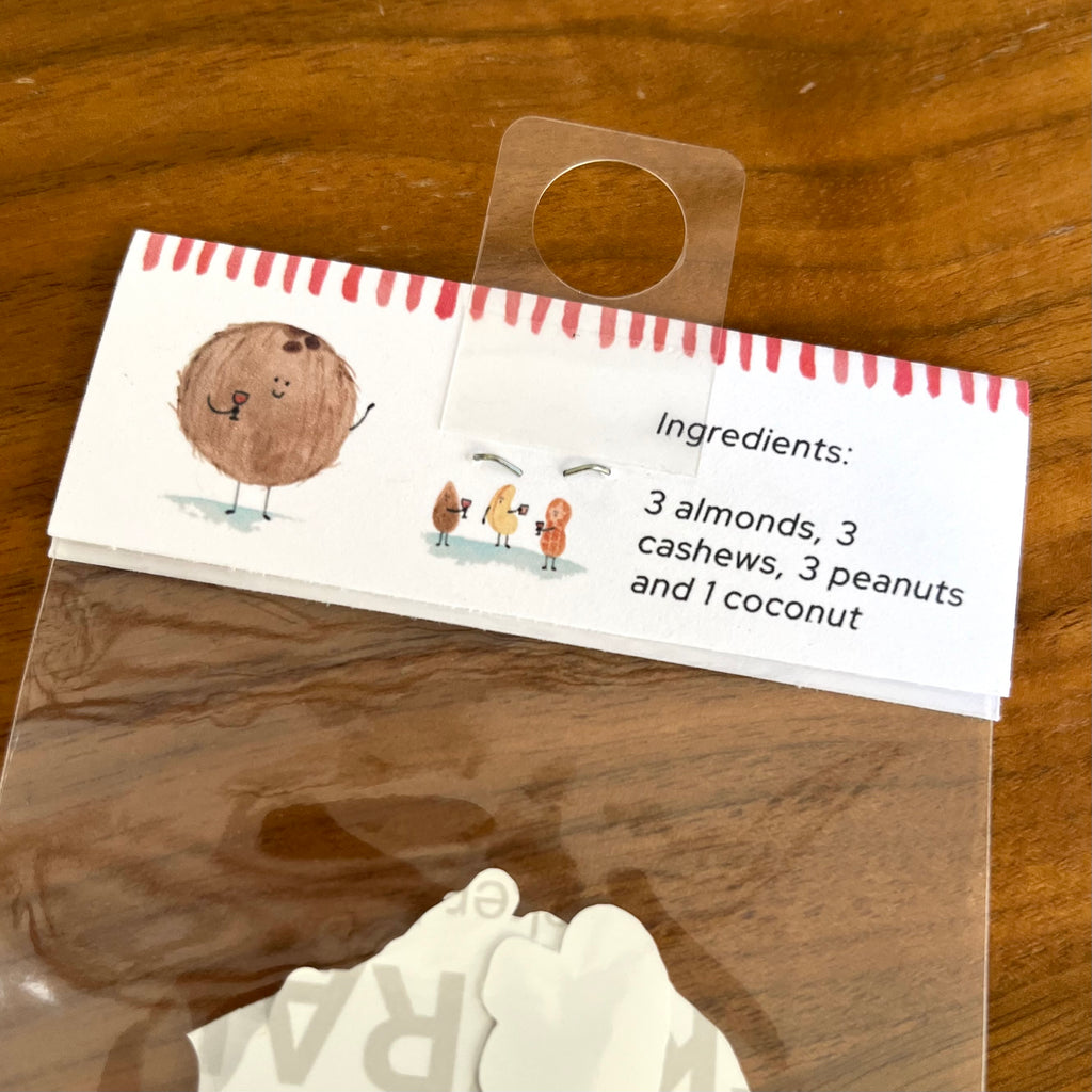 A packaged set of white coconut-shaped decorative pieces inside a clear plastic bag, with a printed paper header card showing a cartoon coconut and the text 'Ingredients: 3 almonds, 3 cashews, 3 peanuts and 1 coconut' on a wooden surface.