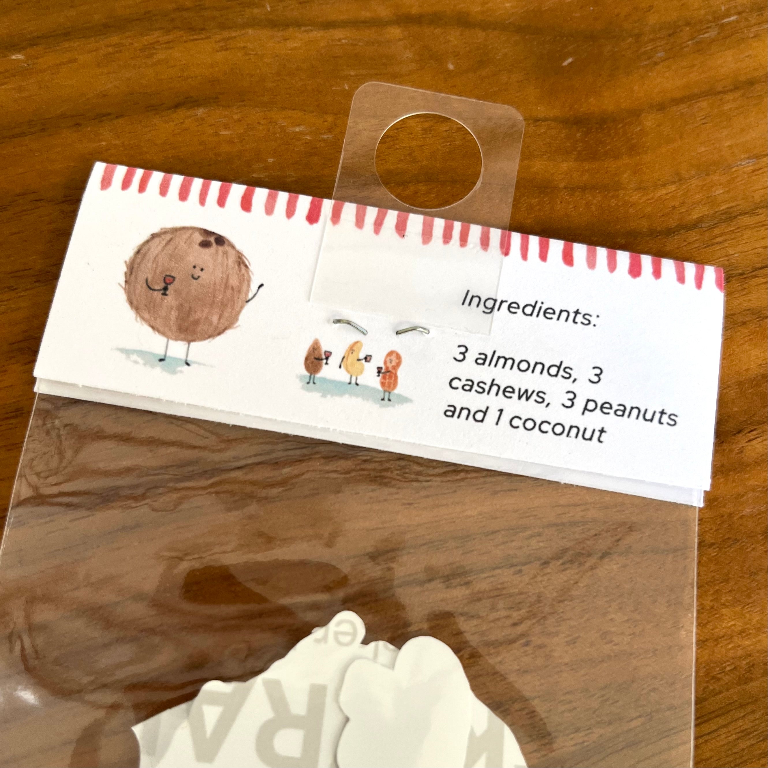 A packaged set of white coconut-shaped decorative pieces inside a clear plastic bag, with a printed paper header card showing a cartoon coconut and the text 'Ingredients: 3 almonds, 3 cashews, 3 peanuts and 1 coconut' on a wooden surface.
