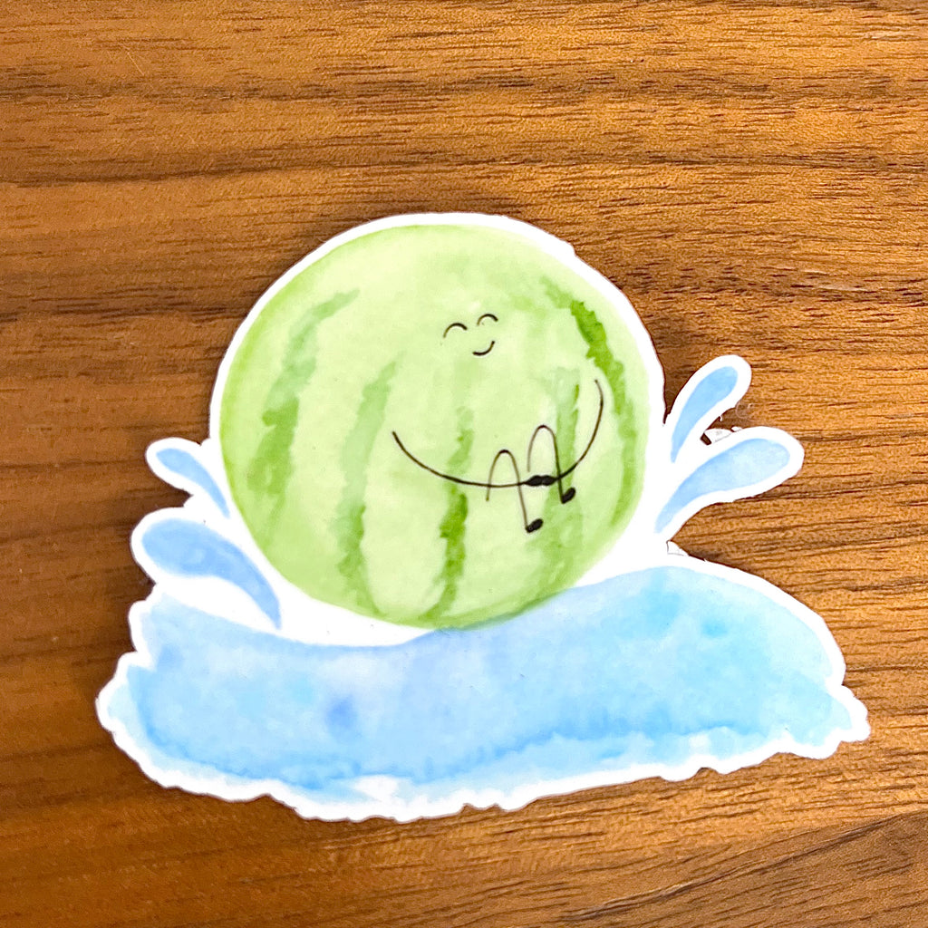A die-cut sticker featuring a cute watercolor illustration of a smiling green watermelon resting on a blue splash, shown on a wooden tabletop.