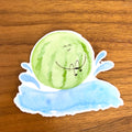 A die-cut sticker featuring a cute watercolor illustration of a smiling green watermelon resting on a blue splash, shown on a wooden tabletop.