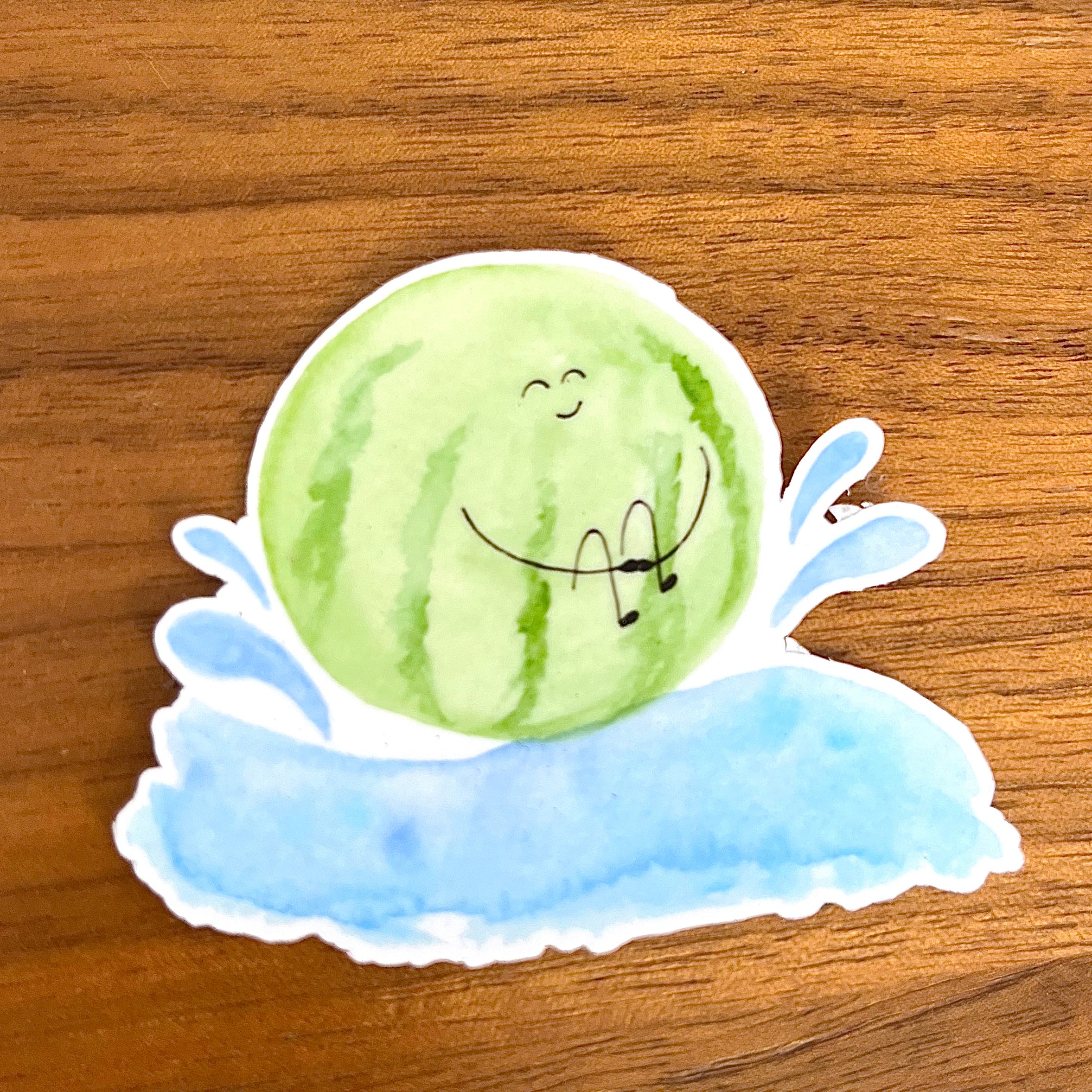 A die-cut sticker featuring a cute watercolor illustration of a smiling green watermelon resting on a blue splash, shown on a wooden tabletop.