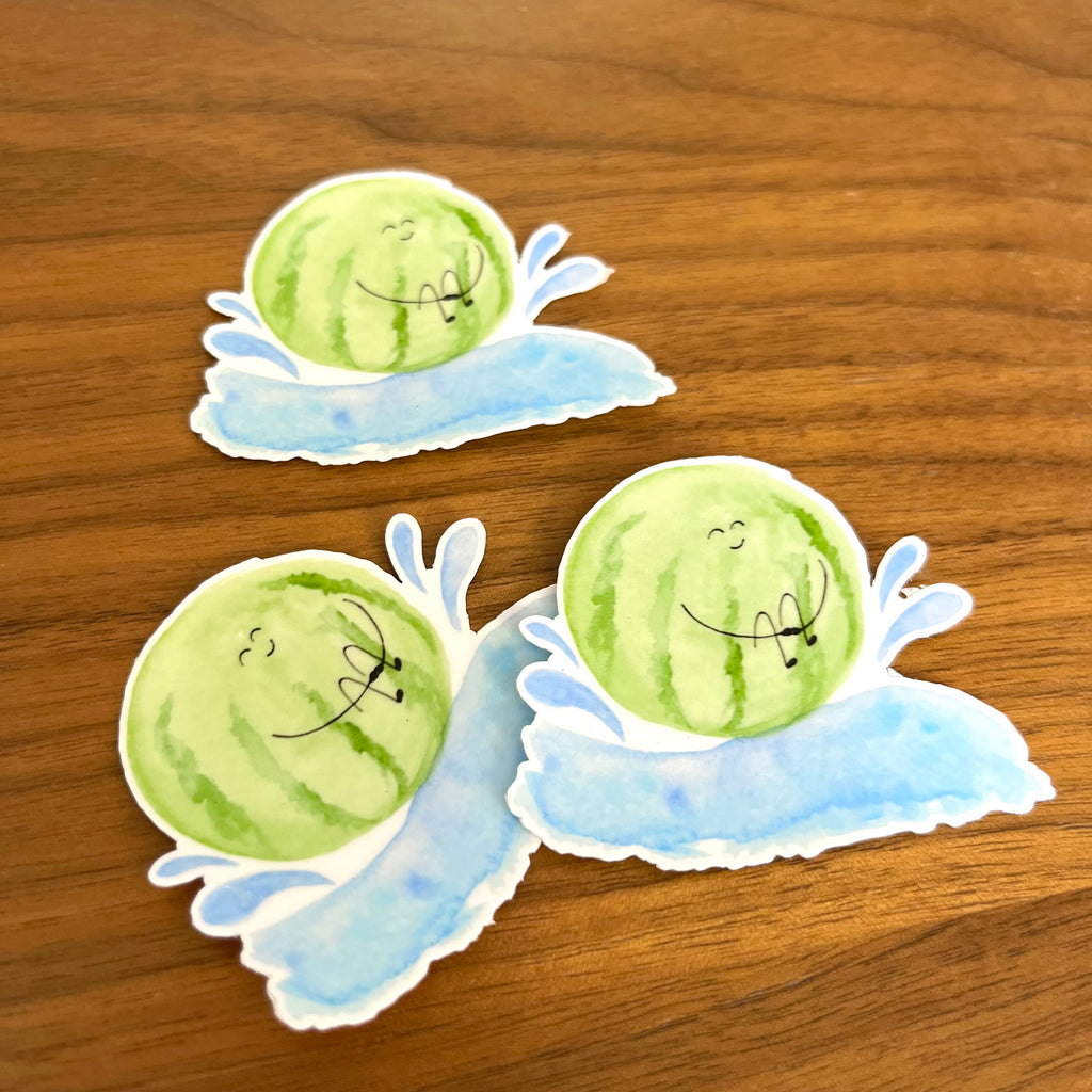 Three die-cut stickers featuring cartoon watermelons with sleeping faces on light blue water splash backgrounds, displayed on a wooden surface.