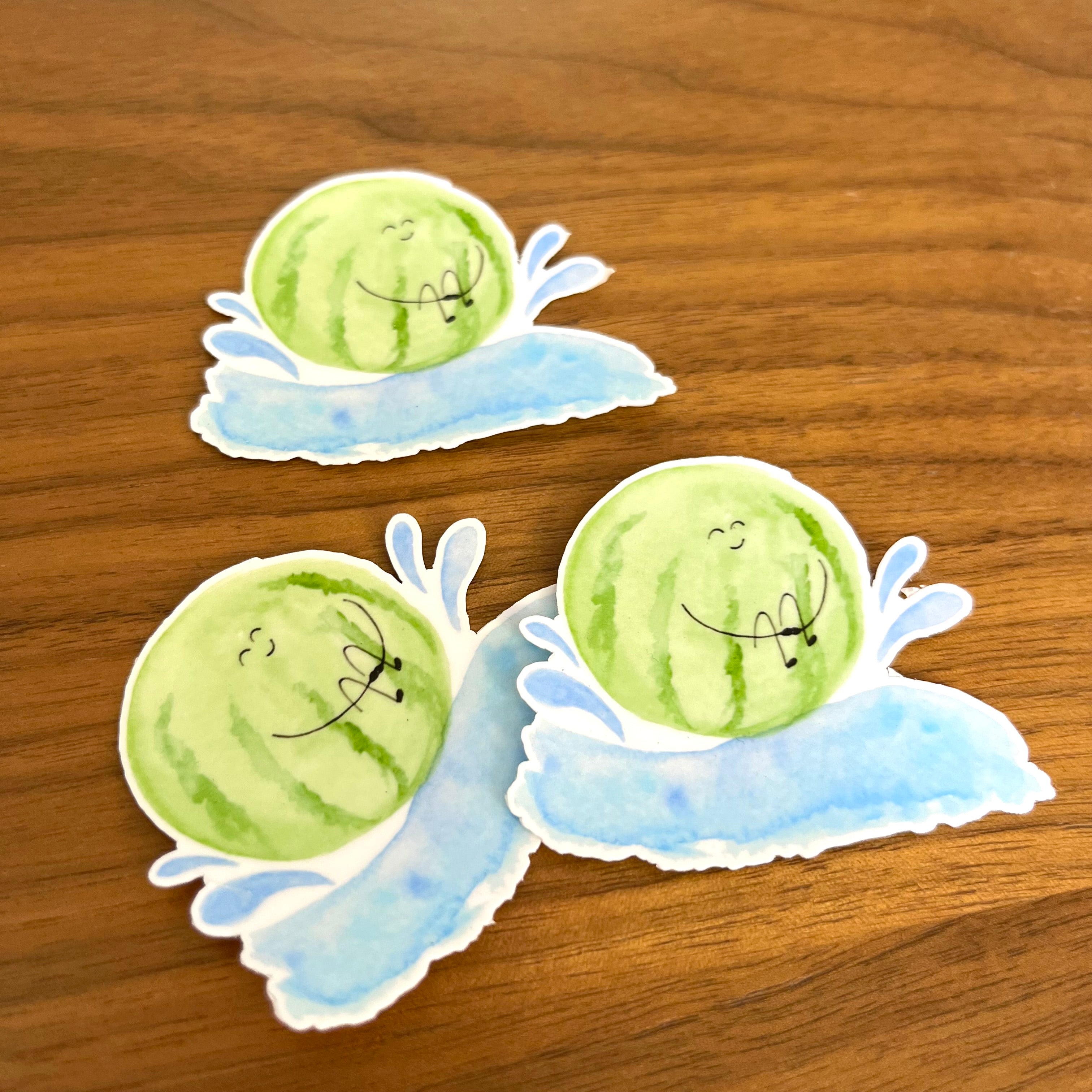 Three die-cut stickers featuring cartoon watermelons with sleeping faces on light blue water splash backgrounds, displayed on a wooden surface.