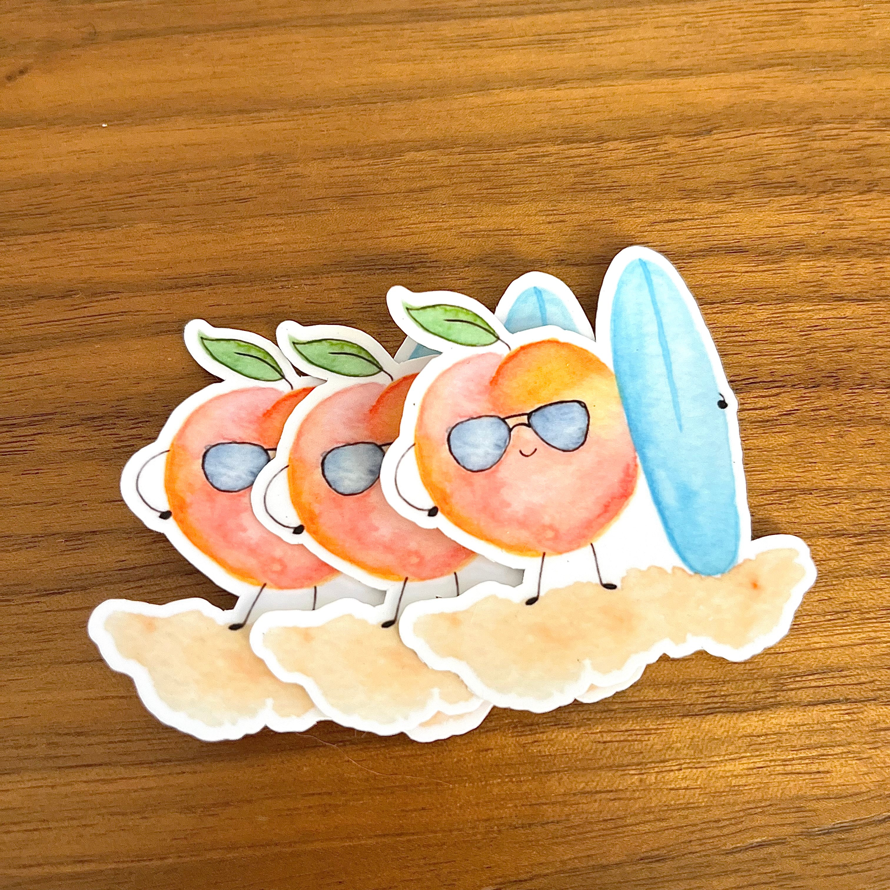 Three cute watercolor peach stickers wearing sunglasses and standing on clouds, with one blue surfboard sticker, arranged on a wooden surface.
