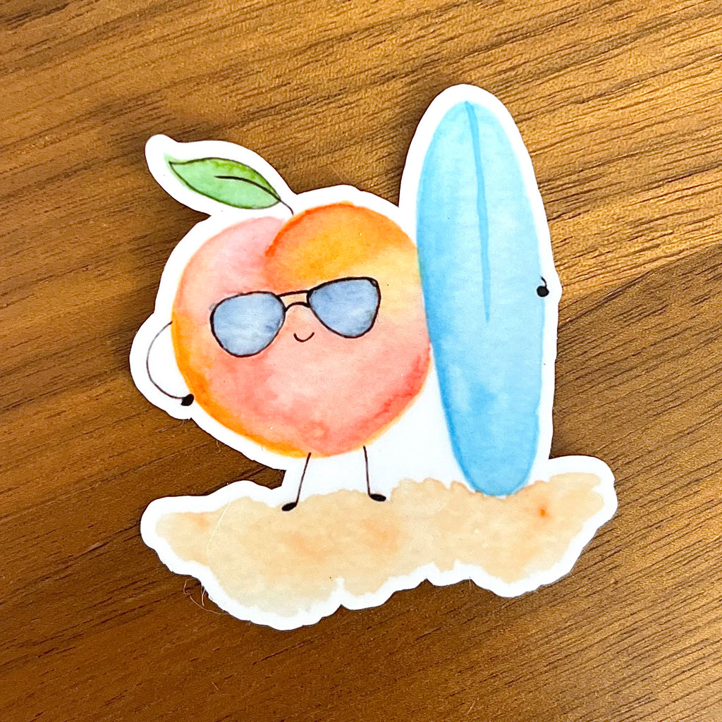 A die-cut sticker featuring a cute peach character wearing blue sunglasses and standing next to a light blue surfboard on a sandy base, placed on a wooden surface. 