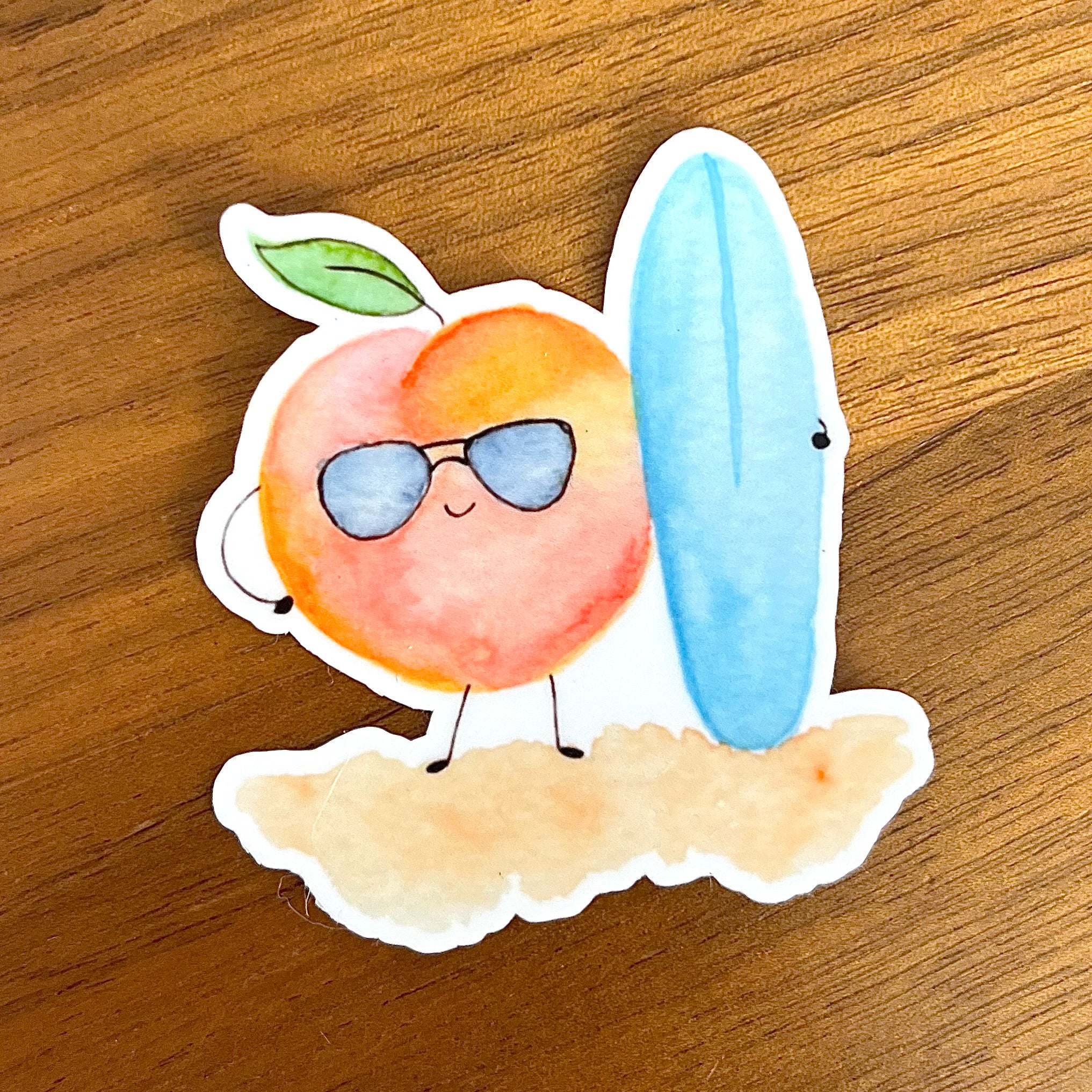 A die-cut sticker featuring a cute peach character wearing blue sunglasses and standing next to a light blue surfboard on a sandy base, placed on a wooden surface. 