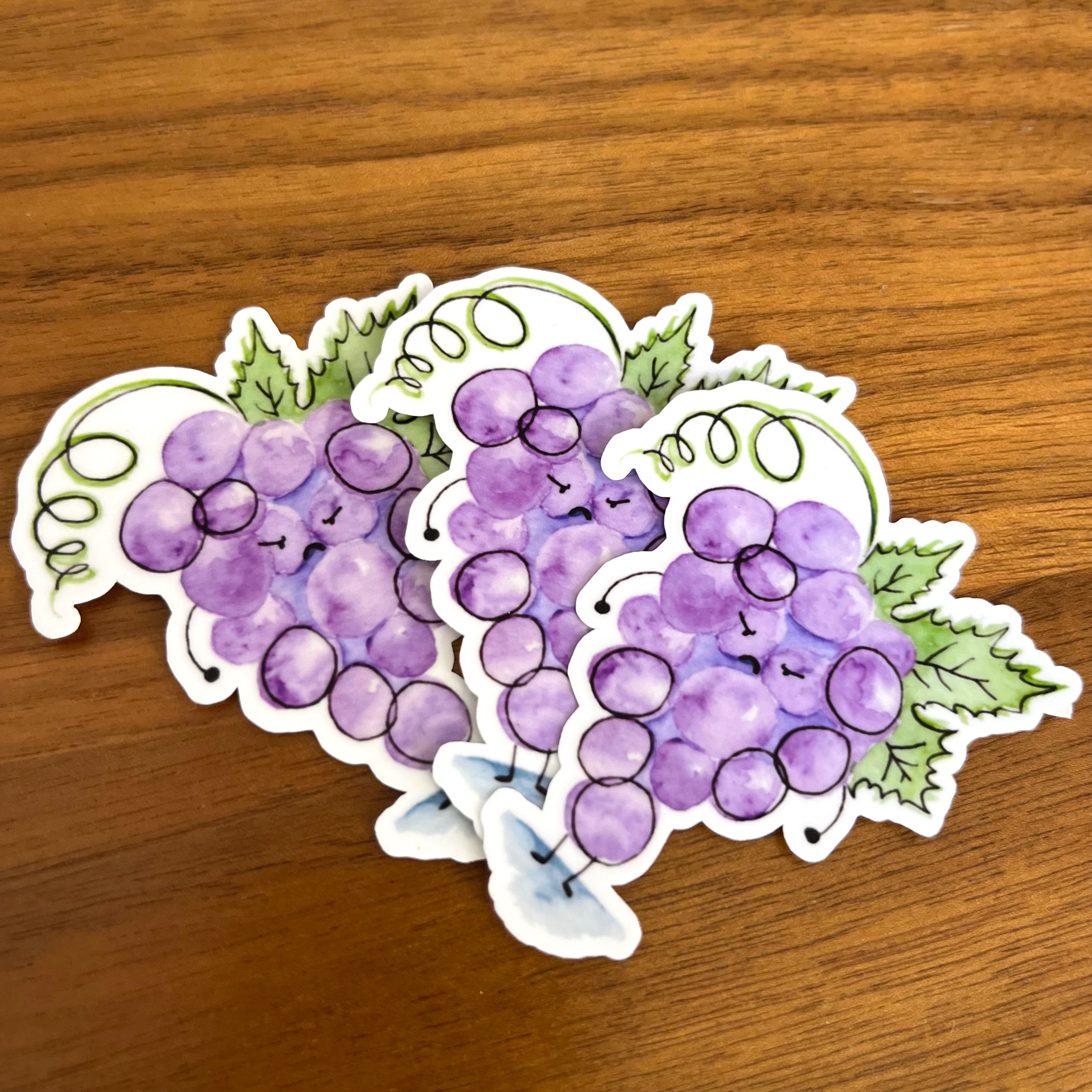 Three die-cut vinyl stickers featuring watercolor-style purple grape clusters with green leaves and vines, arranged on a brown wooden surface.