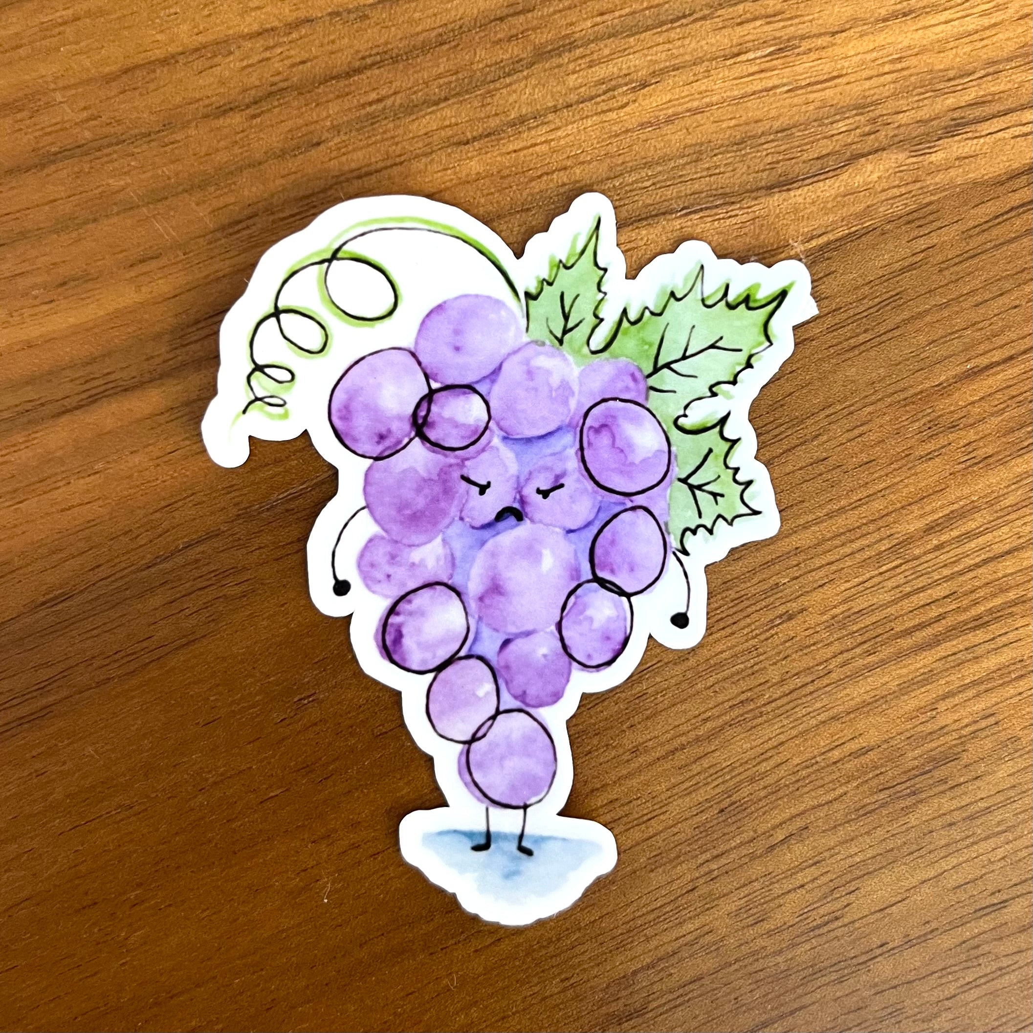A die-cut sticker featuring a cute, watercolor-style illustration of a smiling purple grape bunch with green leaves and a vine, placed on a brown wooden surface.