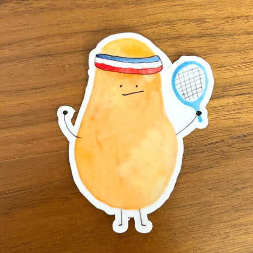 A die-cut sticker featuring a cartoon potato character wearing a red, white, and blue headband and holding a tennis racket, placed on a wooden surface.