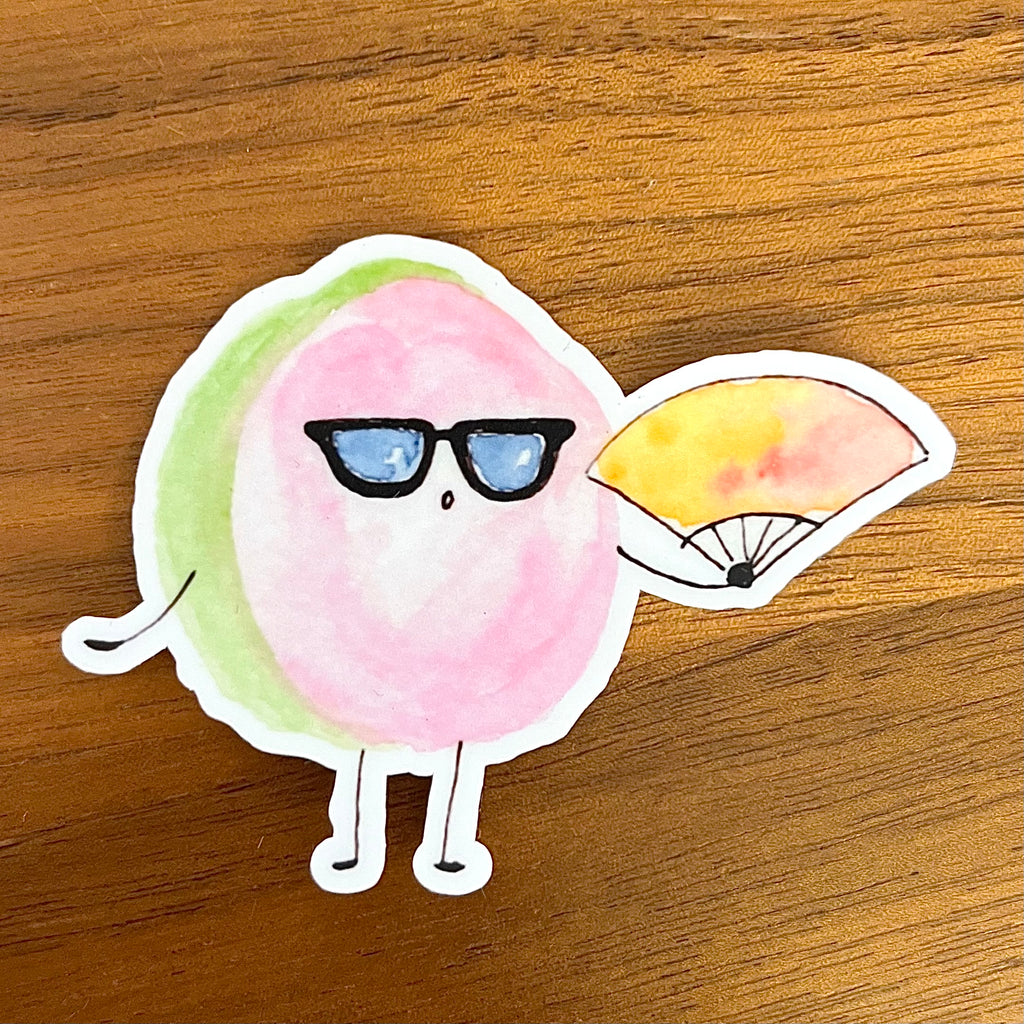 A die-cut sticker featuring a cute cartoon character with a pink body, green side accent, black sunglasses, and a yellow-and-pink fan, placed on a wooden surface.