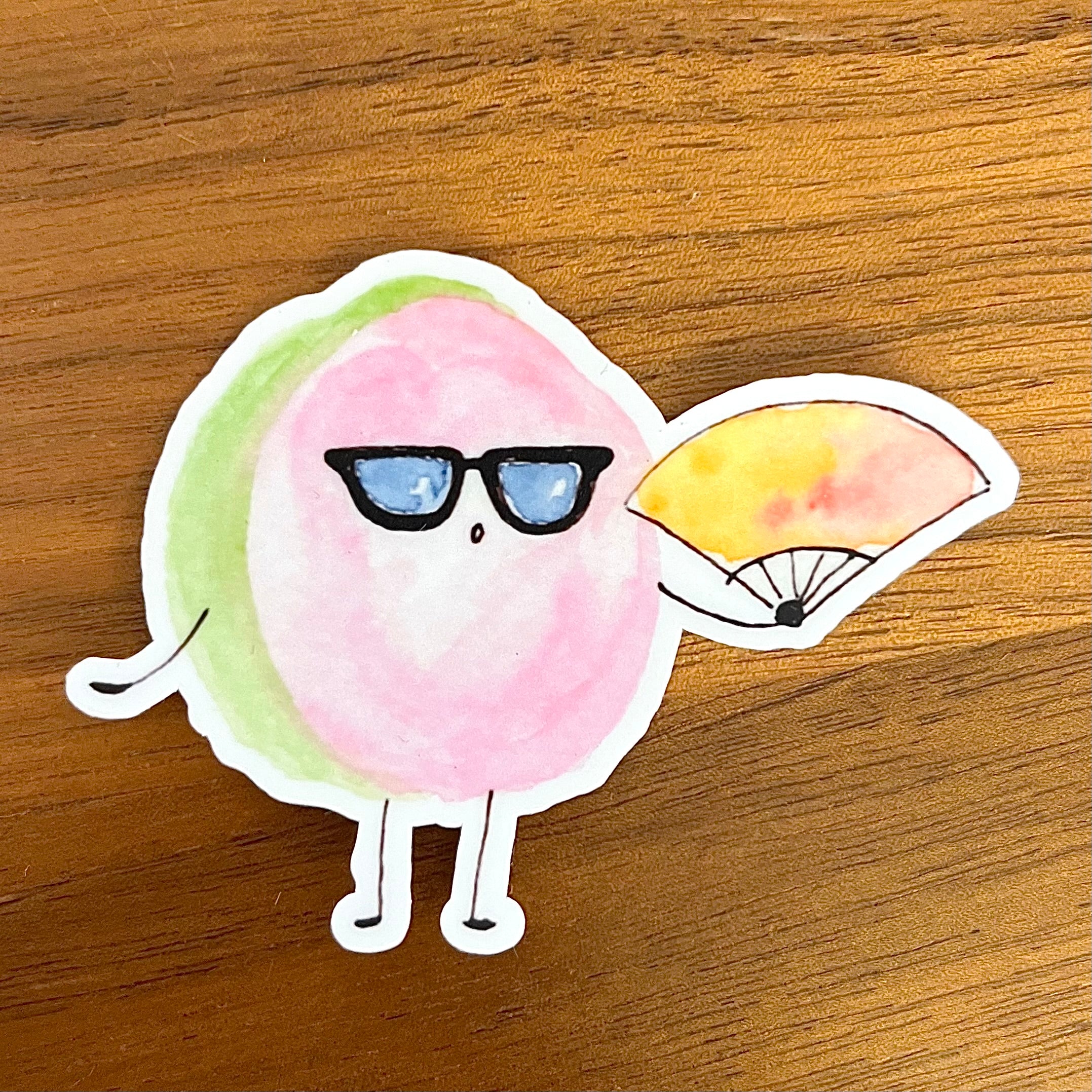 A die-cut sticker featuring a cute cartoon character with a pink body, green side accent, black sunglasses, and a yellow-and-pink fan, placed on a wooden surface.