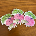 Three cartoon radish stickers with green leafy tops, pink bodies, and purple roots, arranged on a wooden tabletop.