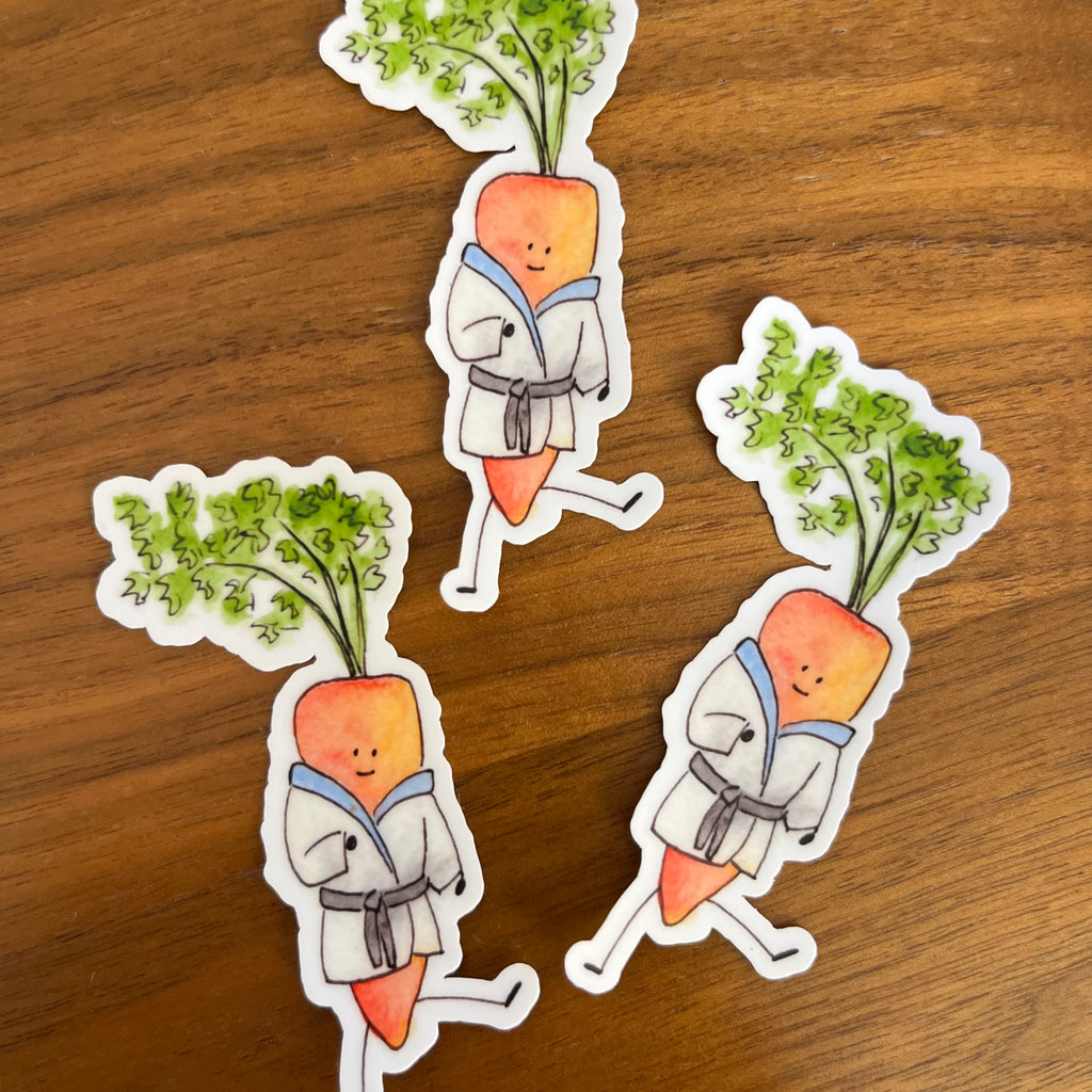 Three die-cut stickers featuring cute illustrated carrots in white karate uniforms with green leafy tops, placed on a brown wooden surface.