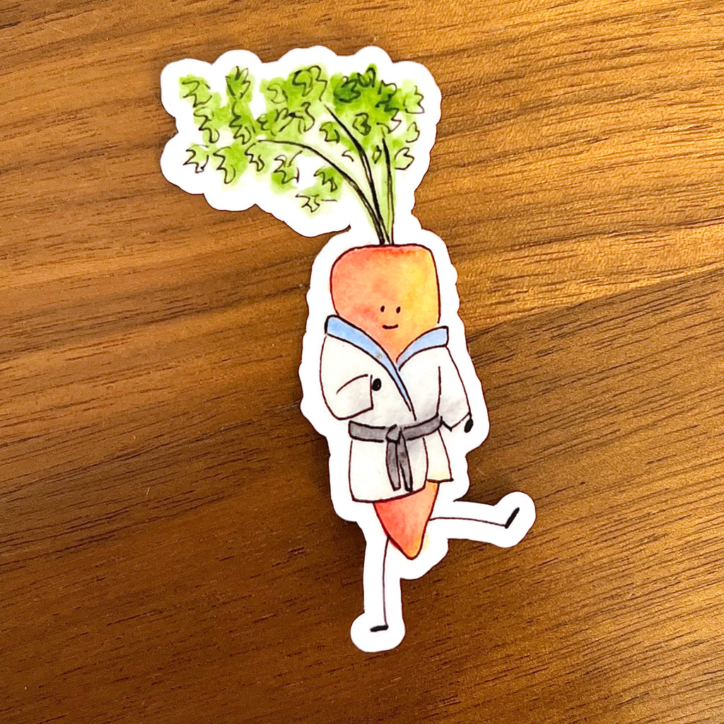 A die-cut sticker featuring a cartoon carrot character wearing a white karate gi with a gray belt and green leafy top, placed on a brown wooden surface.