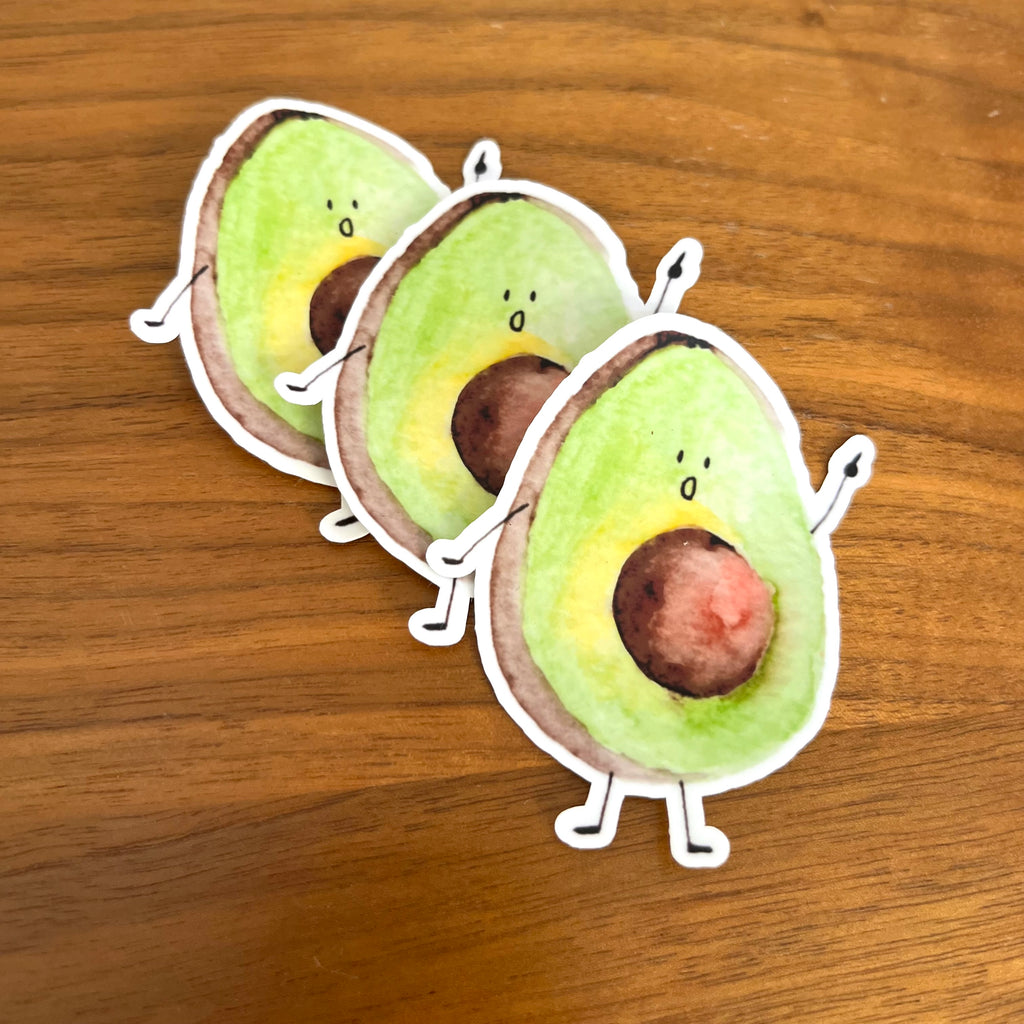 Three die-cut avocado stickers with cute cartoon faces and small arms and legs, arranged diagonally on a wooden surface