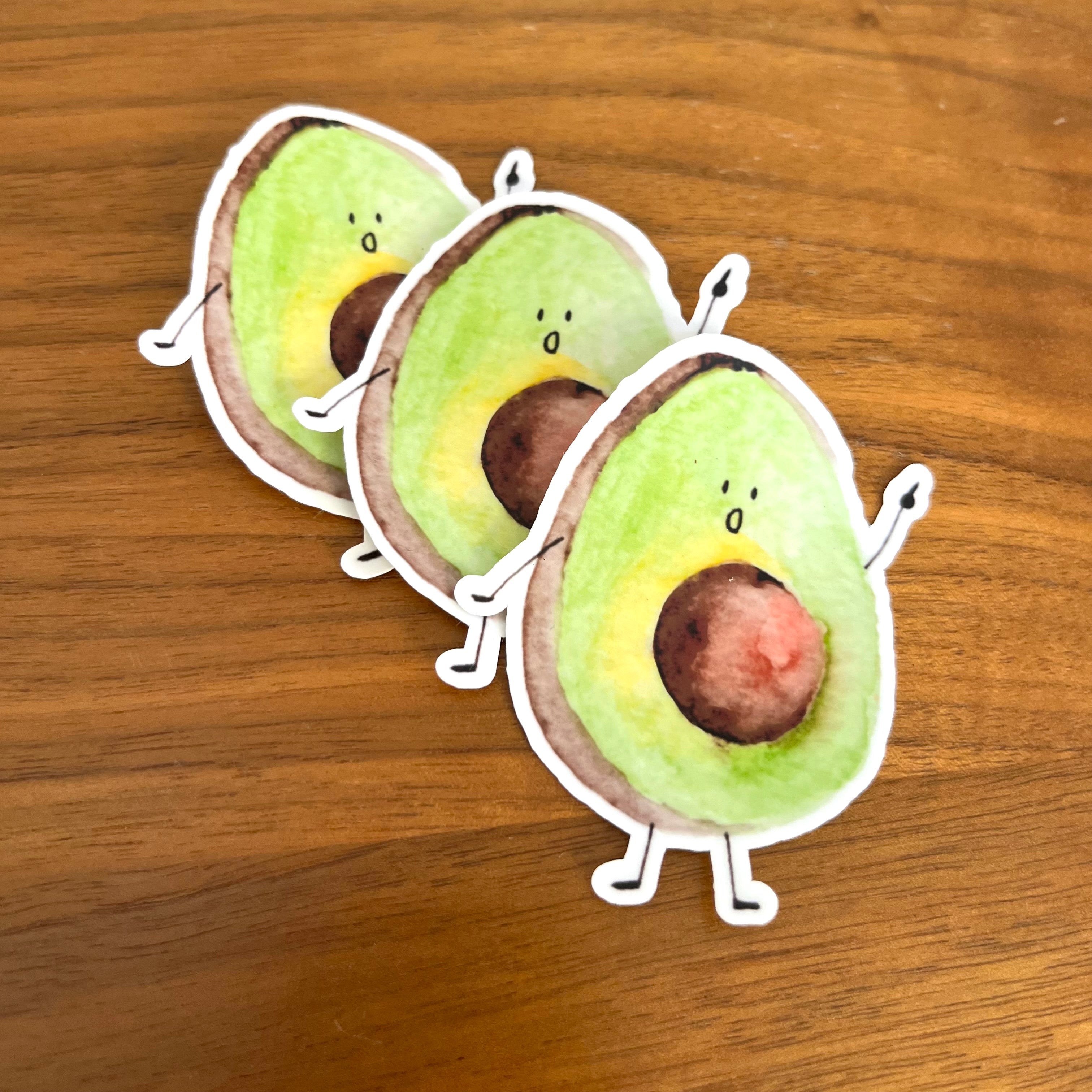 Three die-cut avocado stickers with cute cartoon faces and small arms and legs, arranged diagonally on a wooden surface