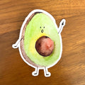 A die-cut sticker of a cute, hand-drawn cartoon avocado with a surprised face, placed on a brown wooden surface