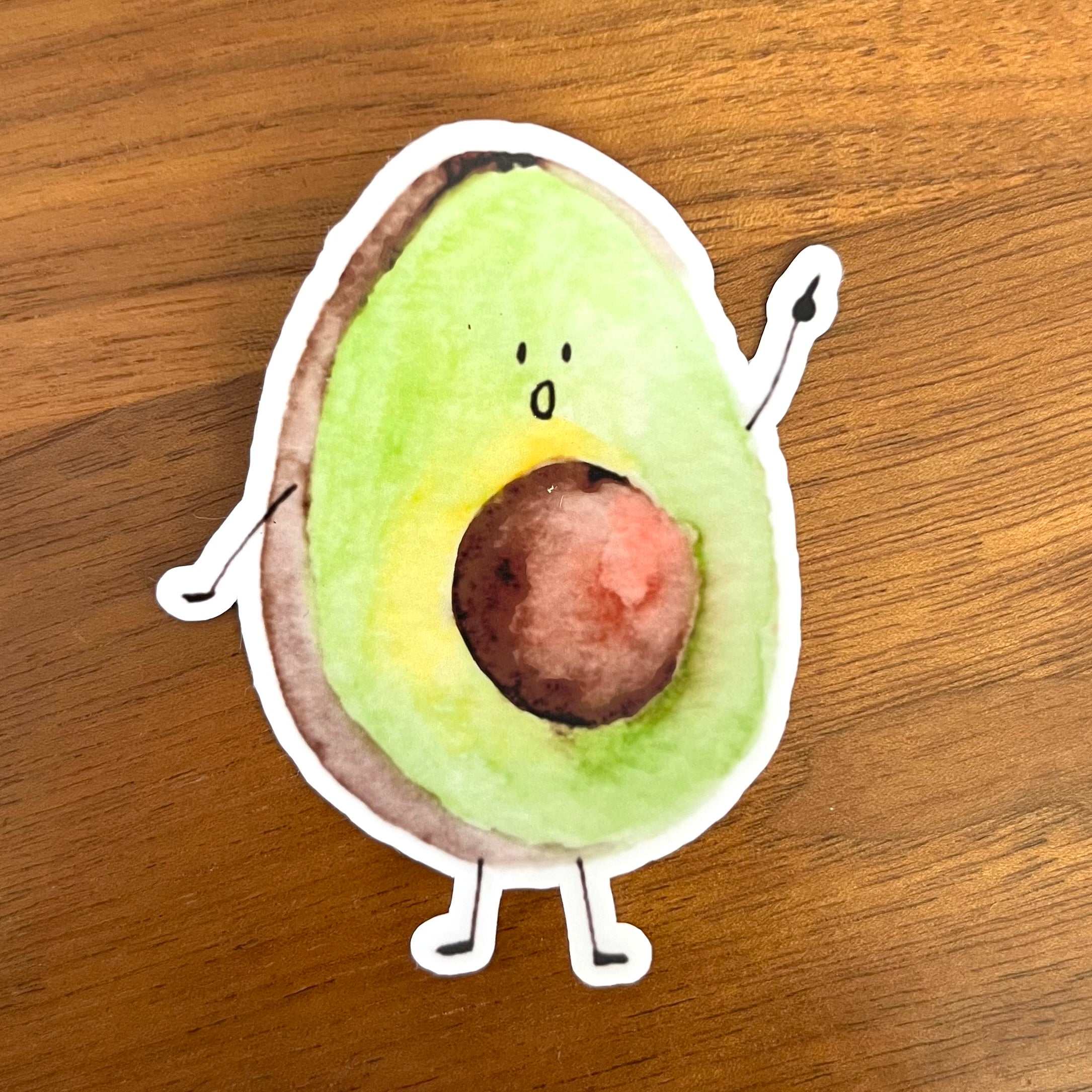 A die-cut sticker of a cute, hand-drawn cartoon avocado with a surprised face, placed on a brown wooden surface