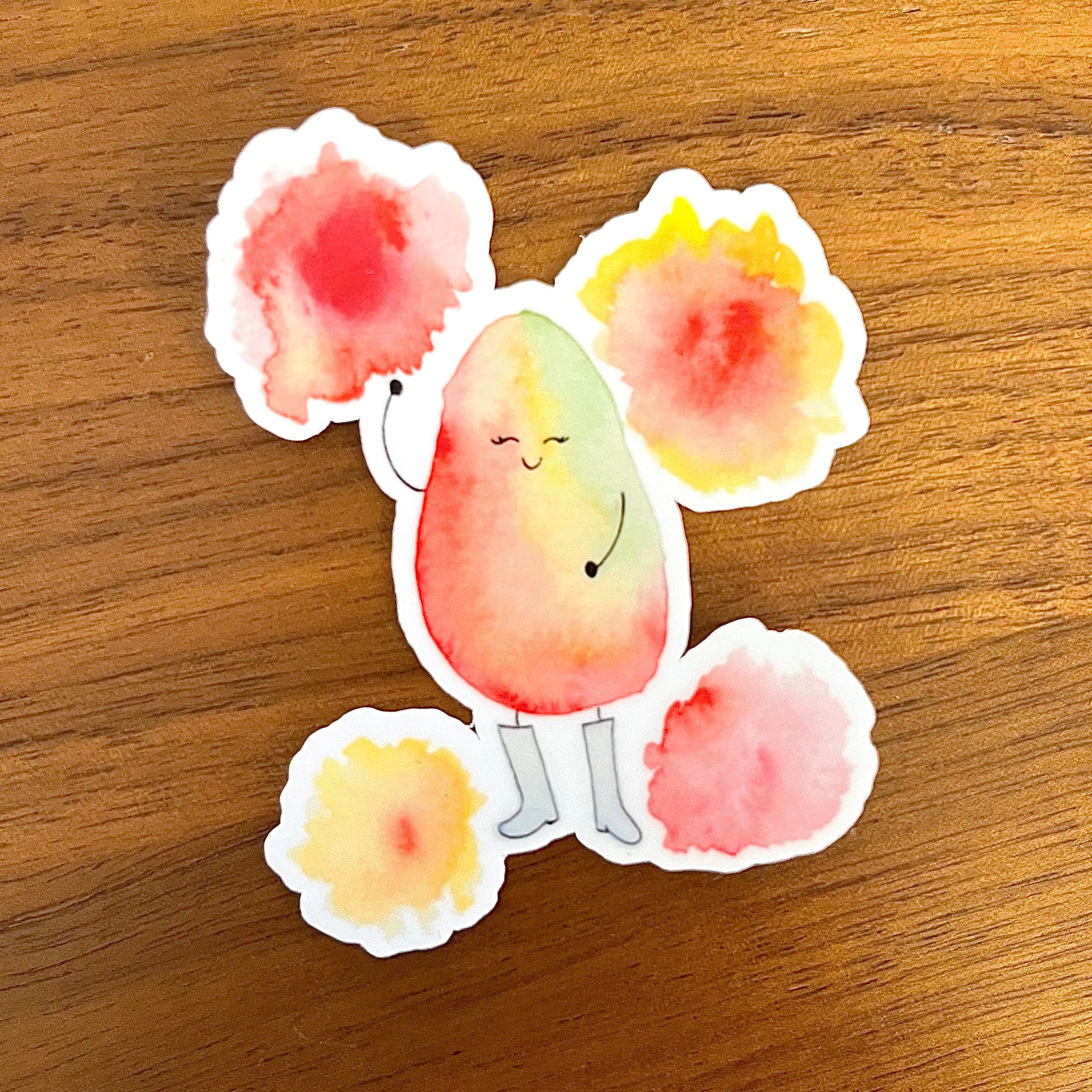 A die-cut sticker featuring a cute watercolor-style character with a peach and yellow body, pink flower-like heads, and gray boots, placed on a wooden surface