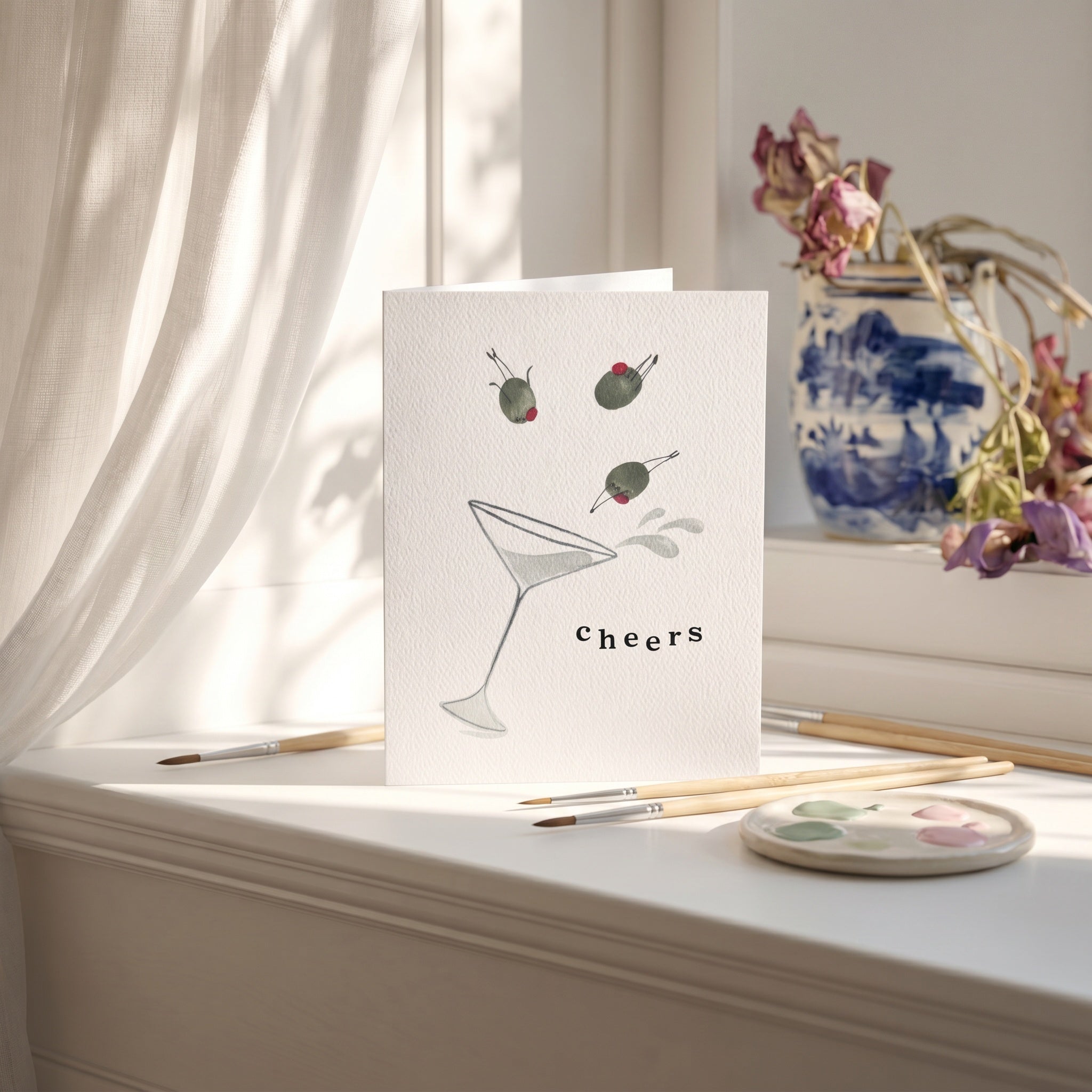 Greeting card with a martini glass and olives design on a white surface with art supplies.