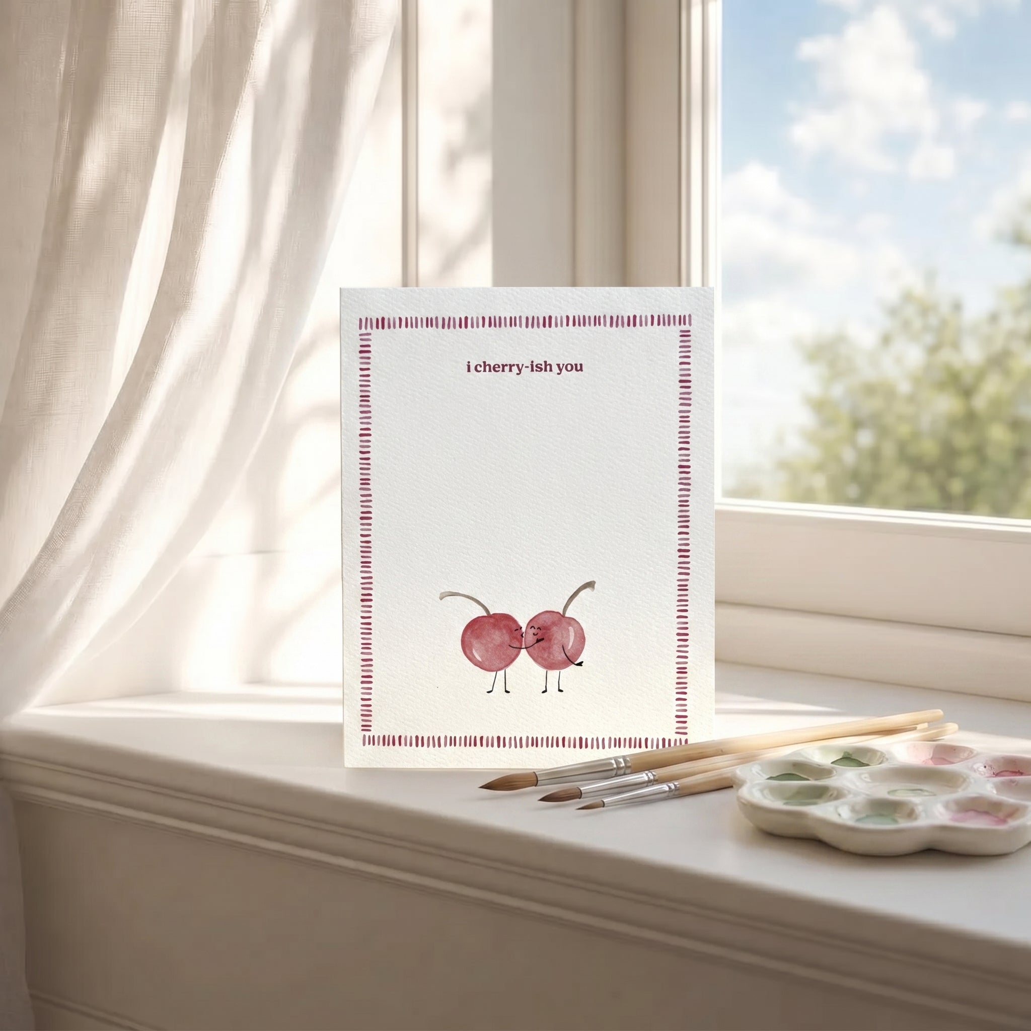 Card with cherry illustration and text on a windowsill with paintbrushes and palette
