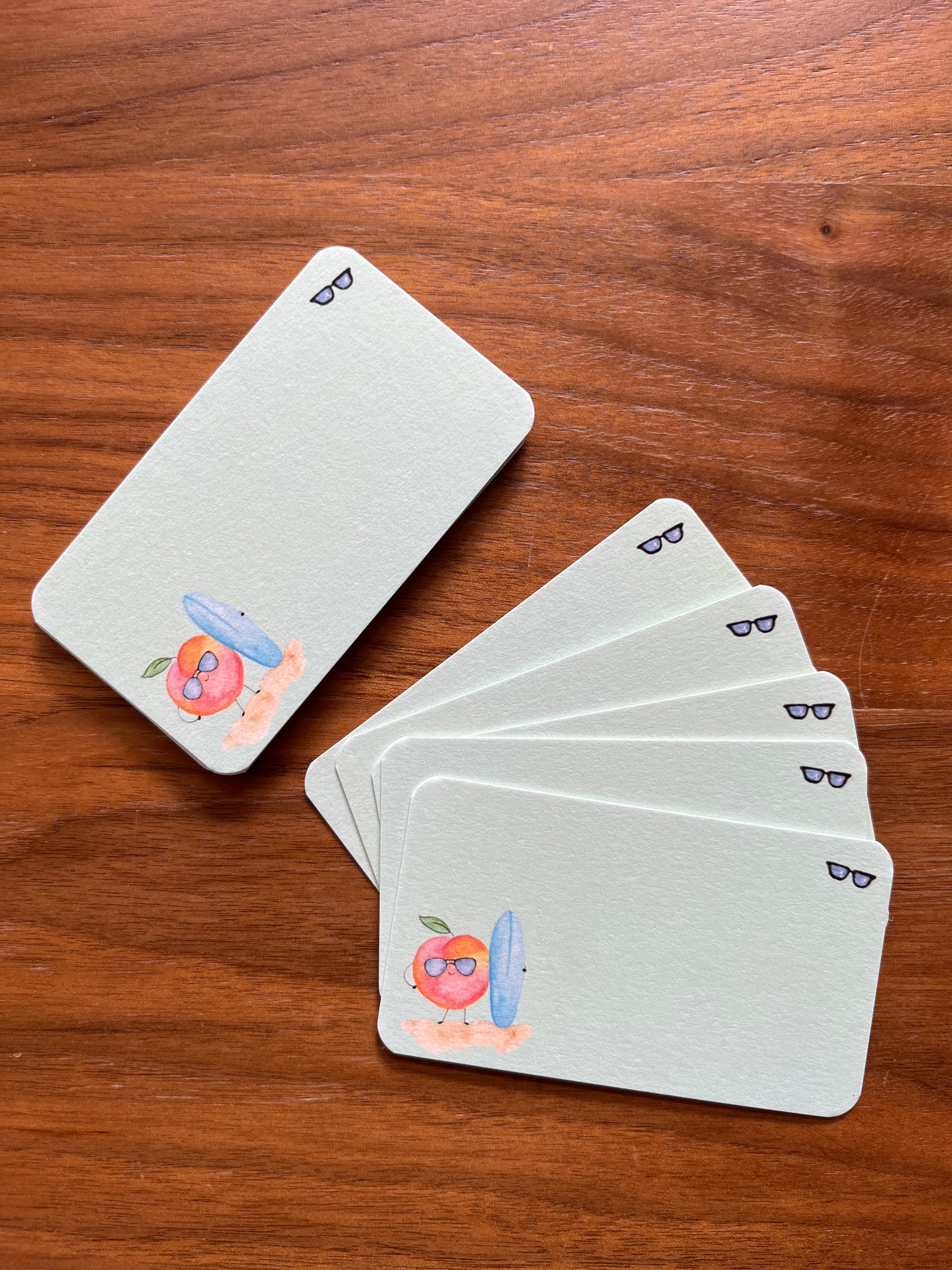 mei pie studio watercolor peach bum mini notes. watercolor peach with a blue sufboard on a green background and sunglasses. printed on classic crest neenah card stock