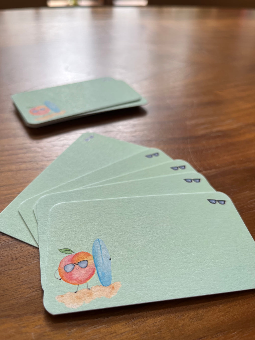 mei pie studio watercolor peach bum mini notes. watercolor peach with a blue sufboard on a green background and sunglasses. printed on classic crest neenah card stock