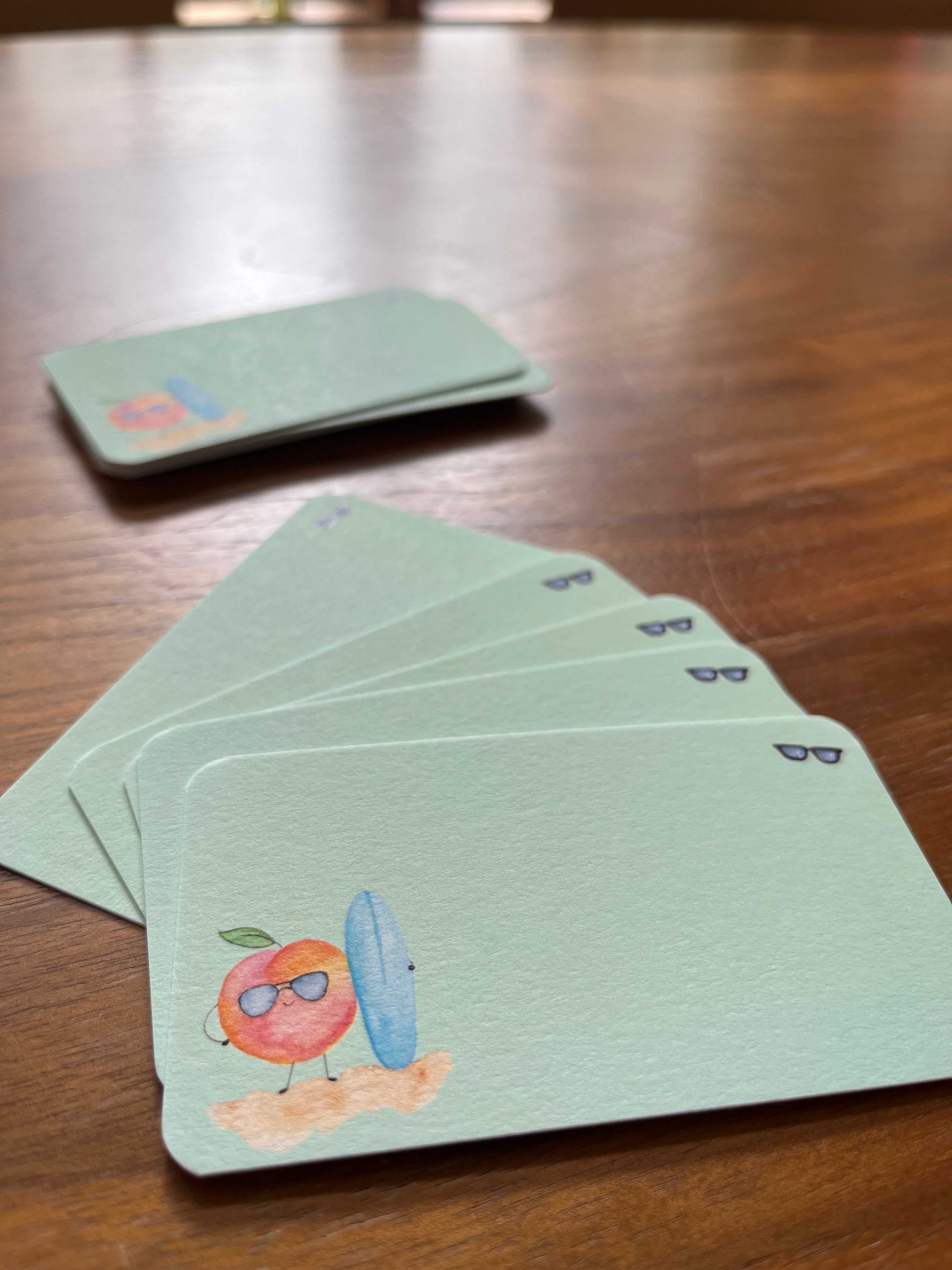 mei pie studio watercolor peach bum mini notes. watercolor peach with a blue sufboard on a green background and sunglasses. printed on classic crest neenah card stock